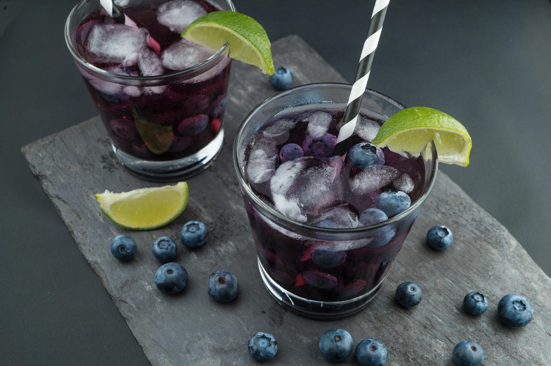 Two clear tumblers filled with a blackcurrant cordial and ice. The glasses are decorated with lime segments and each has a stripey straw. On the slate there are scatted fresh blackcurrants and a slice of lime. Via Unsplash 2017 (artist unknown)