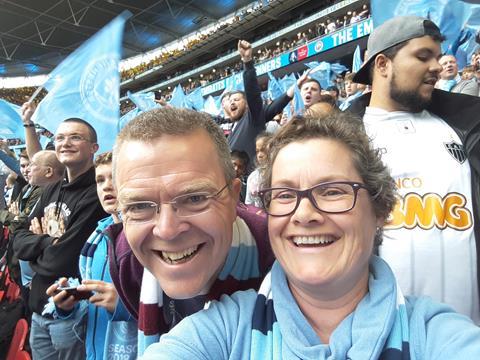 A selfie of two adults wearing the pale blue of Manchester City and smiling broadly at the camera. They are attending the FA Cup final in 2019 at Wembley stadium. Behind them are City fans celebrating and waving their blue flags.