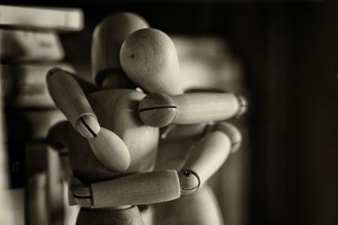 A blak and white photograph of two wooden artist mannequins in a hug position. In the background are some blurry images of books. By Marco Bianchetti via Unsplash
