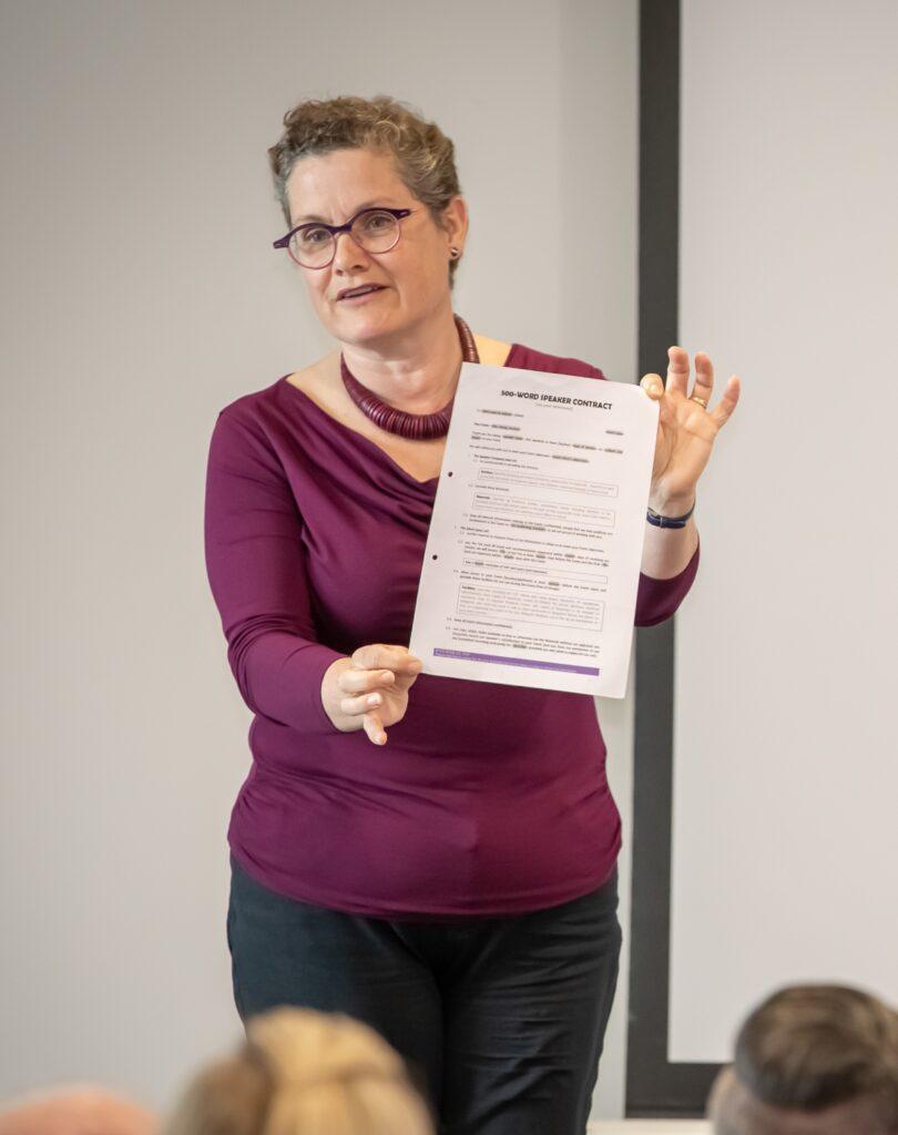 A lady wearing a berry-coloured top and holding a single page (500-word) speaker contract in front of her